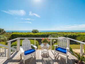 Una terraza de madera con dos sillas Adirondack blancas con cojines azules, una mesita redonda con superficie de vidrio y una maceta con flores rojas.