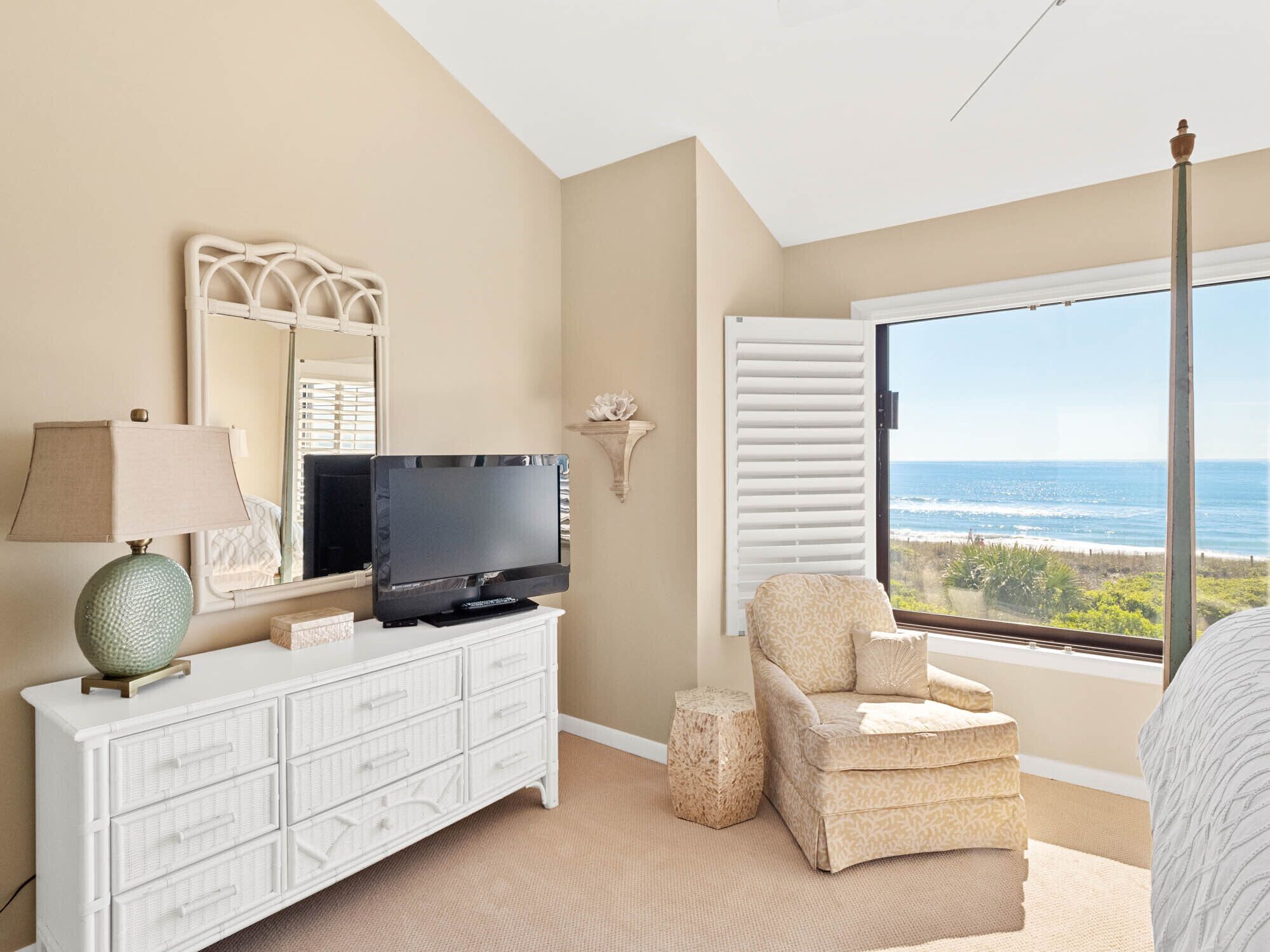Ein Schlafzimmer mit Fernseher auf einer weißen Kommode, einem Sessel, einem kleinen runden Hocker, einem großen Fenster mit Meerblick und einem Deckenventilator.