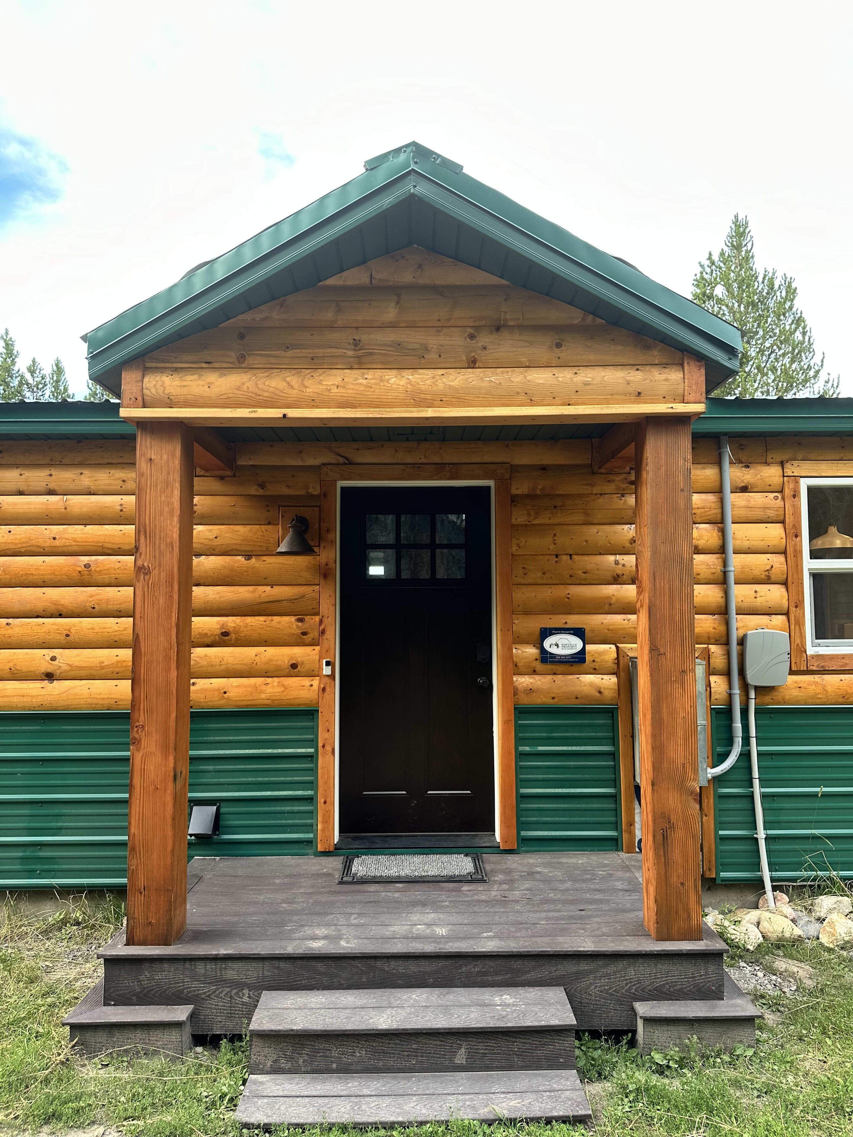 A log cabin with a green metal roof, a wooden porch, and a black door.