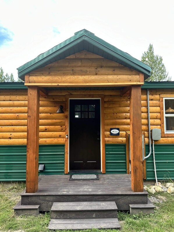 A log cabin with a green metal roof, a wooden porch, and a black door.