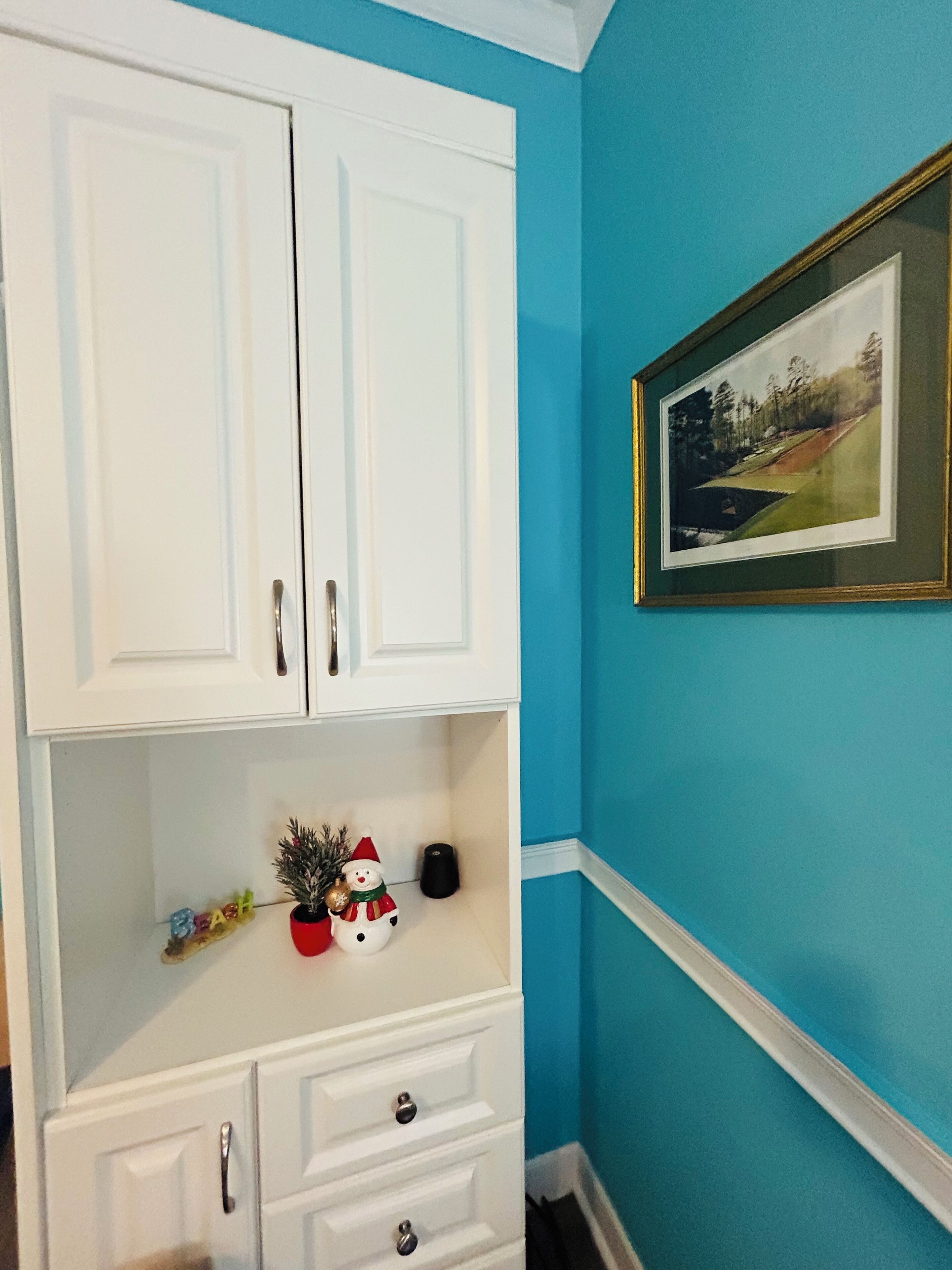A white cabinet with drawers and a shelf, a framed picture on the wall, and a small Christmas decoration.