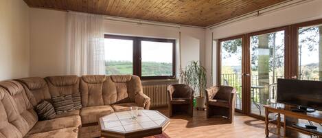 Una sala de estar con un sofá modular grande, una mesita de centro y un televisor sobre un mueble de madera. Hay dos sillones, una planta en maceta y vistas al exterior a través de amplios ventanales.