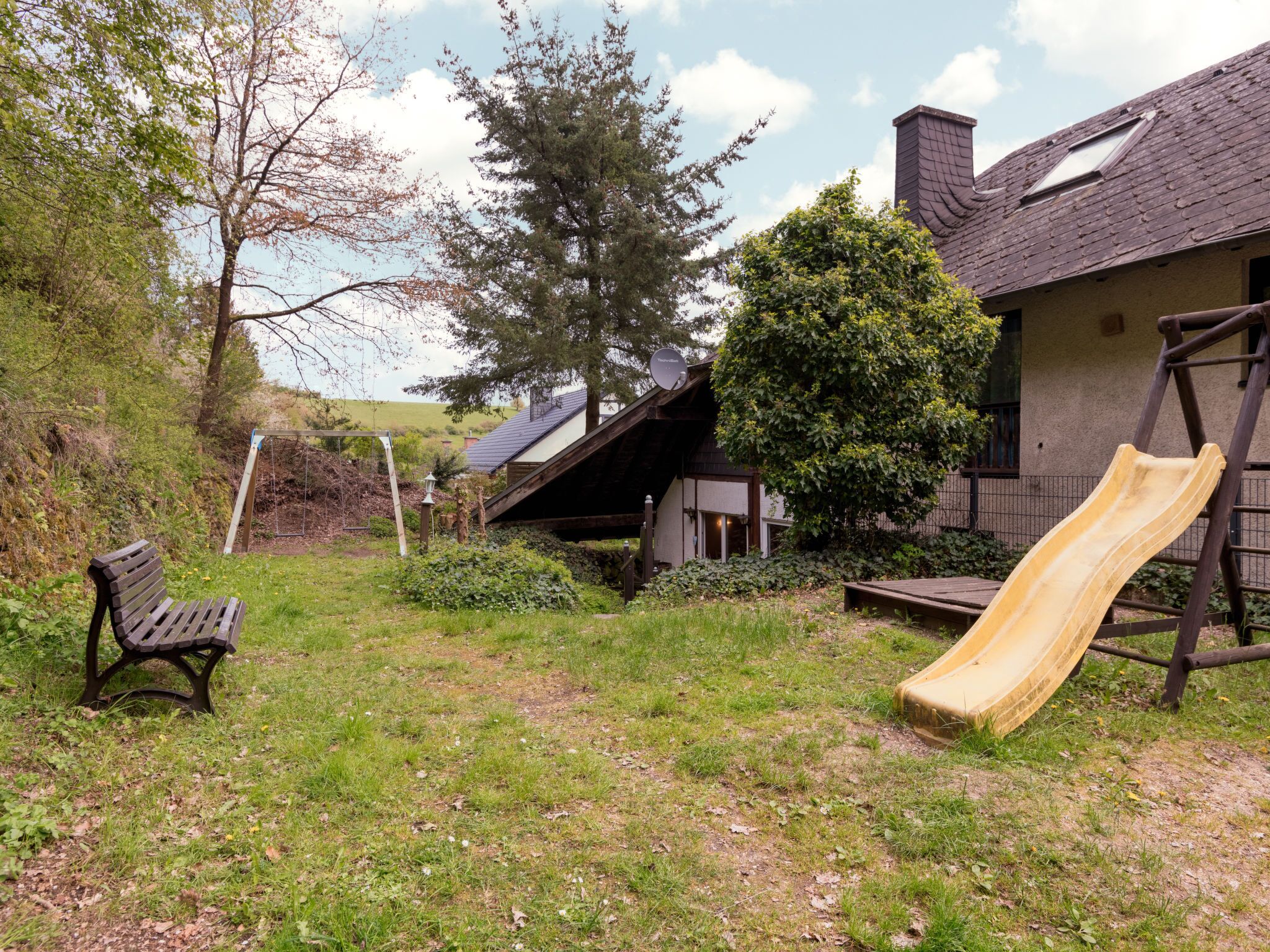 A backyard with a wooden bench, a slide, and a house with a sloped roof.