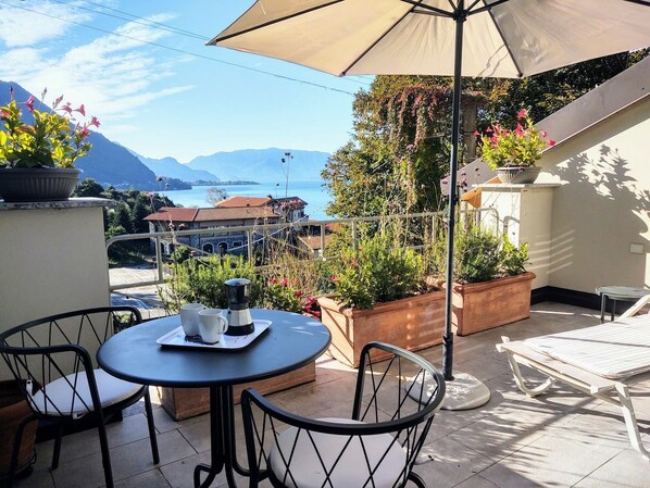 A terrace with a table, chairs, and an umbrella. There are potted plants and a view of mountains and a body of water.