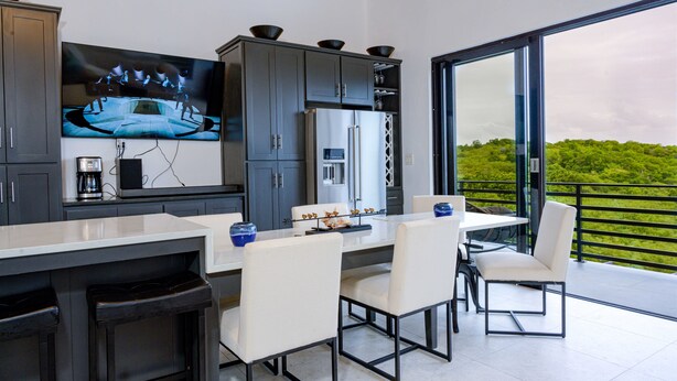 A modern kitchen with a large TV, grey cabinets, and a dining area with white chairs and a glass table. - Carthys Estate (Saint philip)