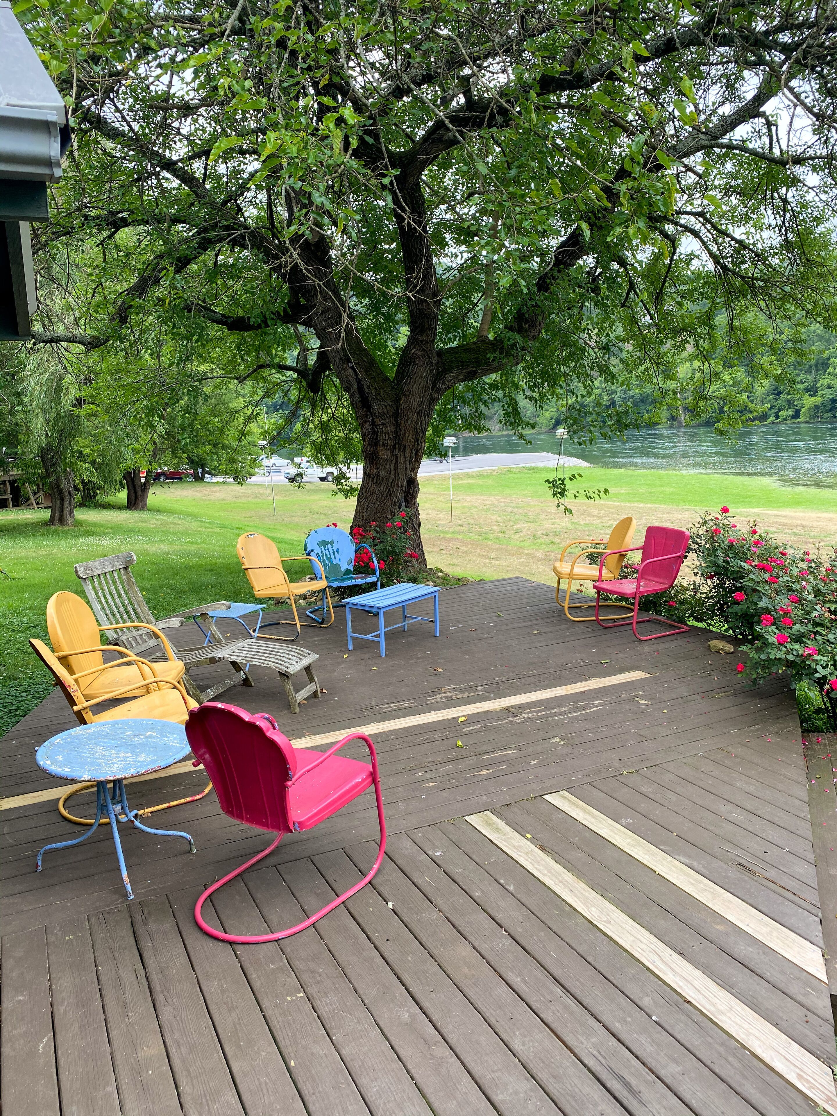 Een houten terras met buitenmeubilair, een grote boom en uitzicht op een rivier.