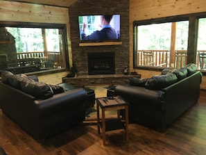 A cozy living room with a stone fireplace, leather sofas, and large windows offering a view of a wooden deck. - Papa Bear’s Retreat, Cedar Falls, 2 BD, 3 BA, Queen Bunk Bed in loft, Fireplaces (Sevierville)