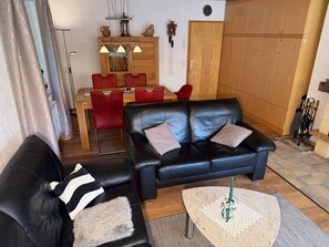 A living room with a black leather sofa, a red dining table with chairs, a wooden cabinet, and a clock on the wall.