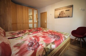 Une chambre avec une armoire en bois, un lit recouvert d’une couvre-lit à motifs floraux, une chaise rouge et un tableau encadré accroché au mur.