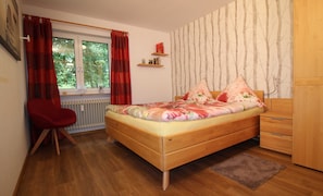 Un dormitorio con cama de madera, una silla roja, radiador, ventana con cortinas, armario y pared con estampado de árbol.