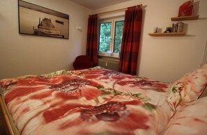 Une chambre à coucher comprenant un lit, une chaise, une fenêtre avec des rideaux, un radiateur et une étagère avec divers objets.