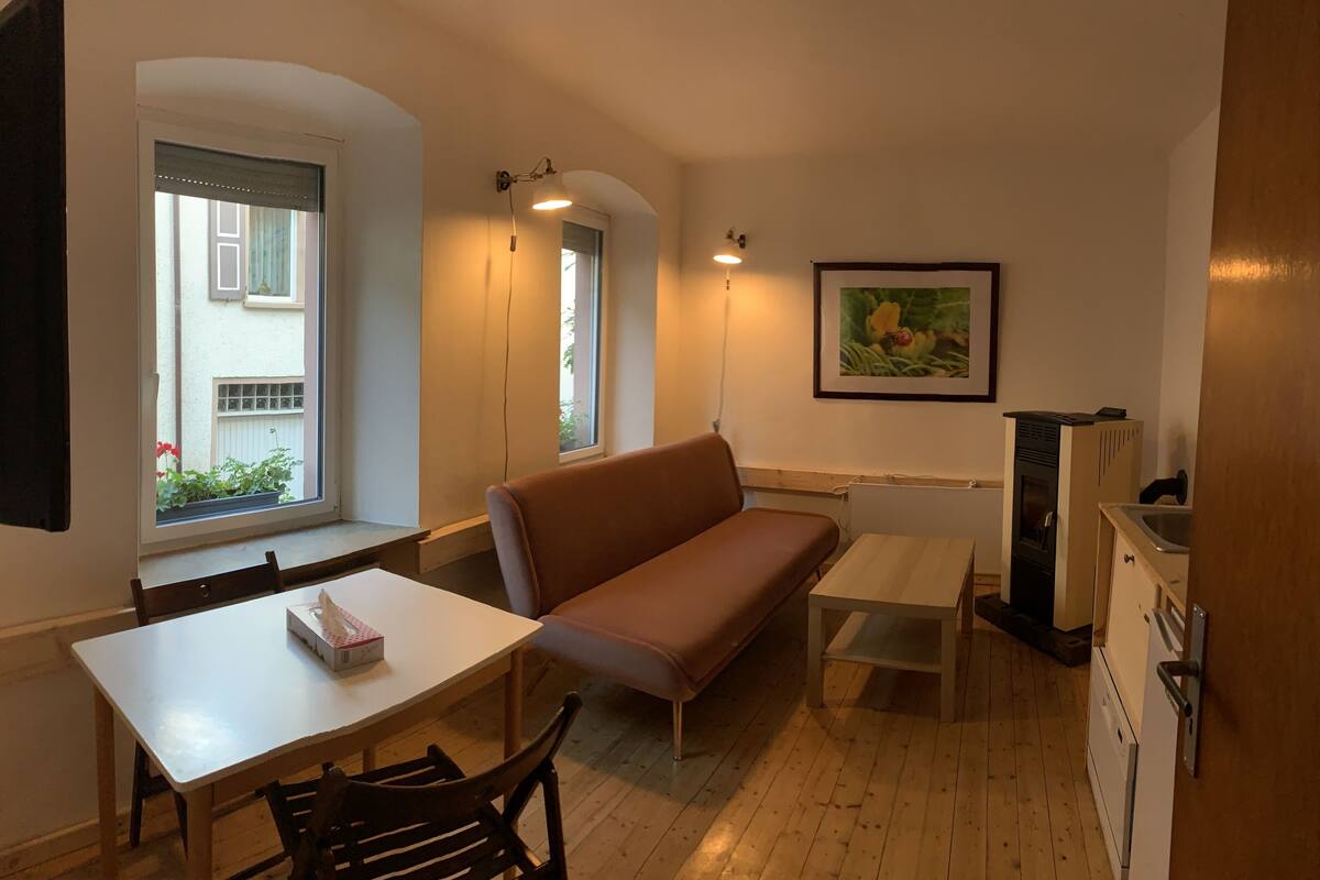 A compact living space with a sofa, a small table, a bench, a TV, and a kitchenette.