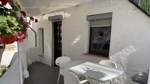 A white exterior of a building with a covered patio area, a white table with chairs, and a window with a blind.