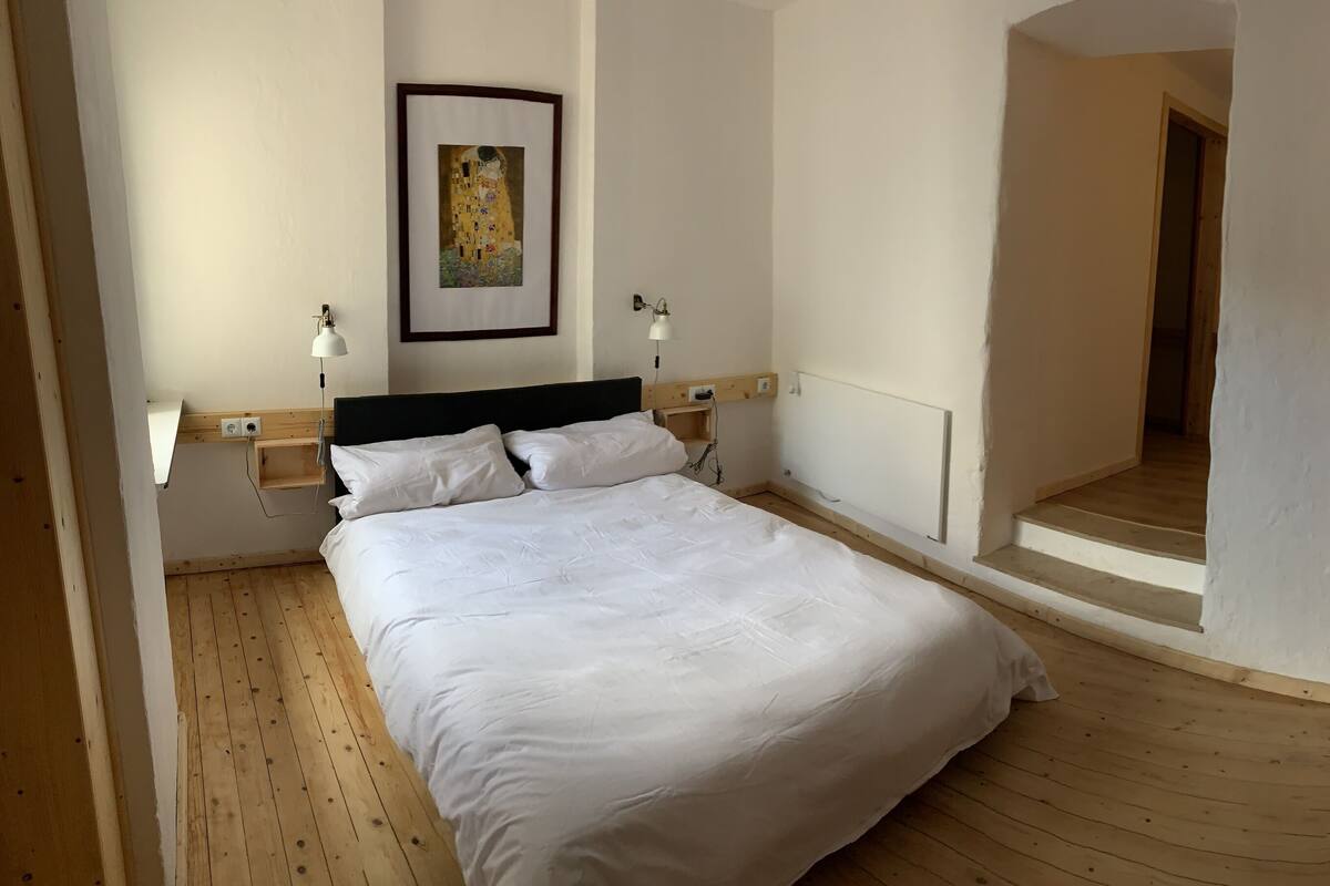 A bedroom with a bed, a nightstand, a lamp, a framed picture on the wall, and a wooden staircase leading to another room.