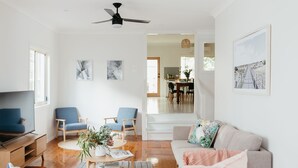 A spacious living room with a flat-screen TV, two blue armchairs, a round coffee table with books and a plant, a beige sofa with a floral throw, and a ceiling fan. - Anchor in Avoca Beach - enjoy your 4 bedroom coastal getaway (Avoca Beach)