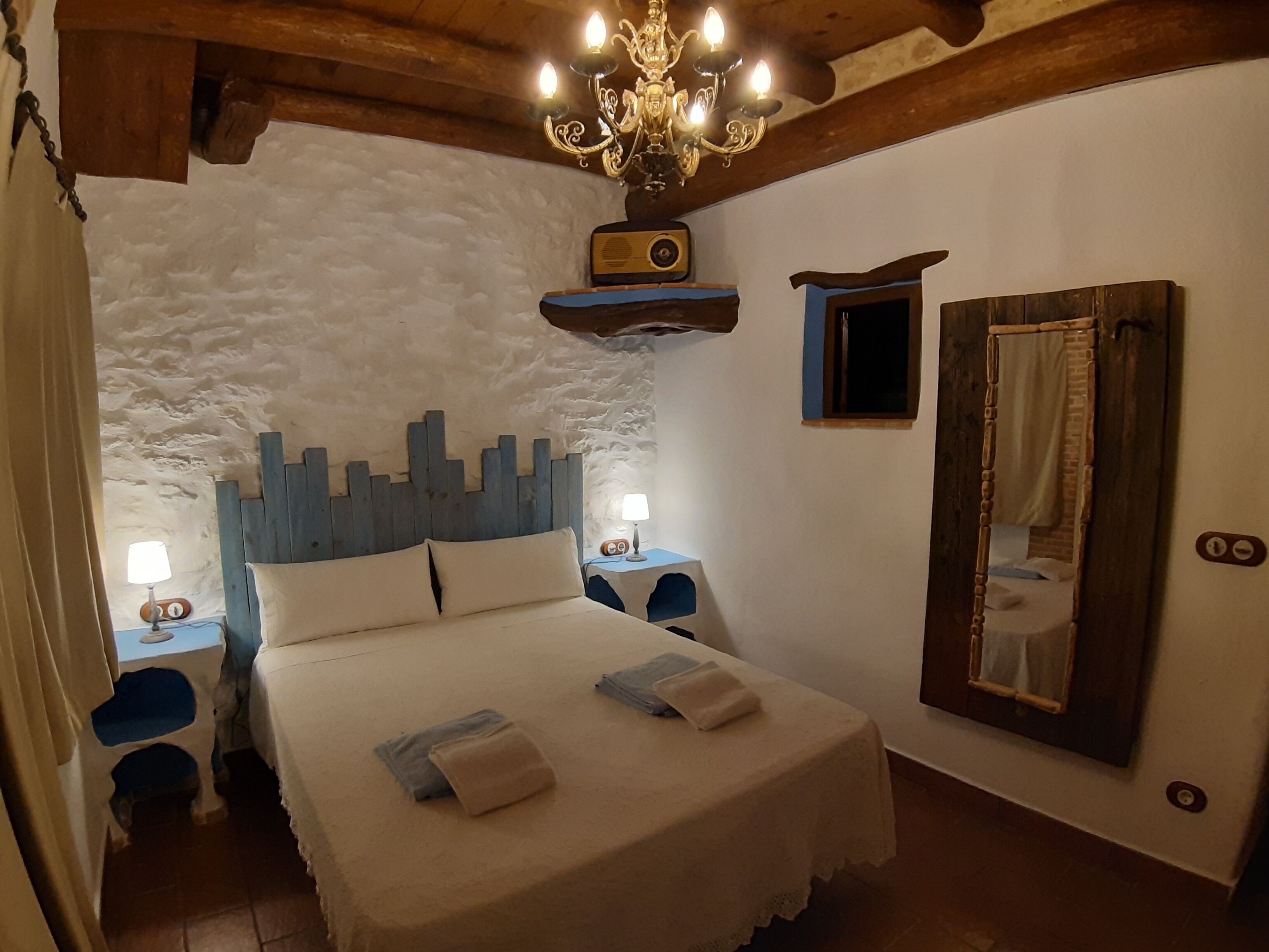 A bedroom with a wooden headboard, a chandelier, a bed with pillows, a nightstand with a lamp, a mirror, and a small shelf with a radio.