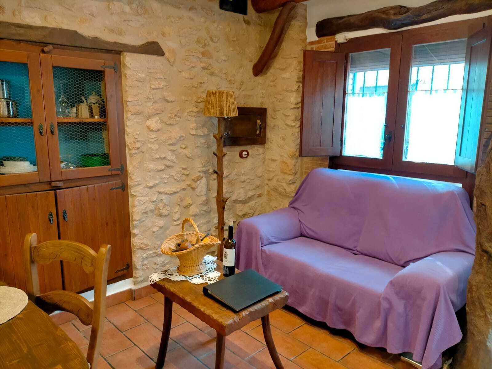 A rustic room with a stone wall, wooden furniture, a purple sofa, a wooden table with a basket, and a window with wooden shutters.