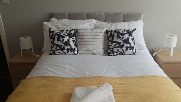 A neatly made bed with a yellow bedspread and a folded white towel, two patterned pillows, and a grey headboard. - Gateshead's Amethyst 3 Bedroom Apt, Sleeps 6 Guest (Chester-le-Street)