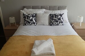 Un lit bien fait, avec une couverture jaune et une serviette blanche pliée, deux coussins à motifs et une tête de lit grise.