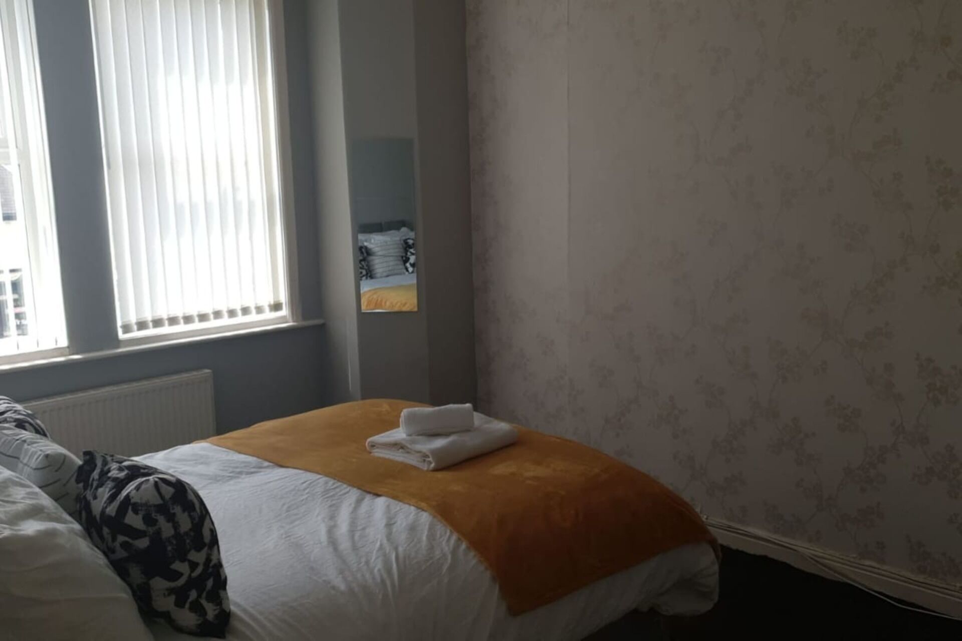 Ein Hotelzimmer mit Bett, einem Fenster mit Jalousien, einem Heizkörper und einer Wand mit floralem Muster.