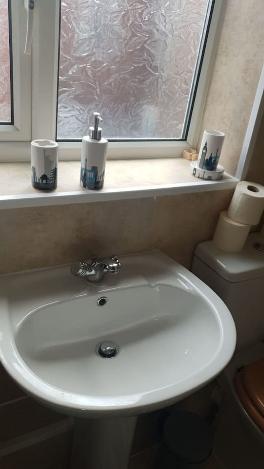 Ein Badezimmer mit einem weißen Waschbecken, einem Fenster mit mattiertem Glas und Toilettenartikeln auf der Fensterbank.