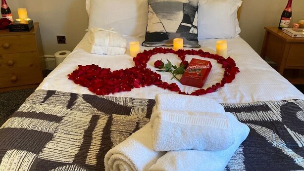 Un lit soigneusement fait, décoré de pétales de rose disposés en forme de cœur, des bougies, une bouteille de champagne et une boîte de chocolats.