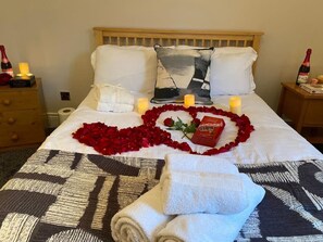 Una cama impecable adornada con pétalos de rosa formando un corazón, velas, una botella de champán y una caja de bombones.