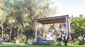 A covered outdoor seating area with a pergola structure, surrounded by lush greenery and trees.