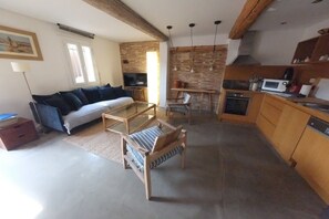 Una sala de estar acogedora con un sofá, una mesa de centro de vidrio y una silla de madera. Hay una zona de cocina con microondas y estufa.