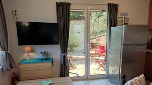 Une pièce compacte équipée d’un téléviseur à écran plat, d’une table en bois, d’un petit canapé, d’un réfrigérateur et d’une porte-fenêtre coulissante donnant sur un espace extérieur avec une table et des chaises.