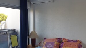 Een slaapkamer met een bed, een raam met gordijnen, een wandairconditioner, een nachtkastje met een lamp en een klein tafeltje met een gele handdoek.