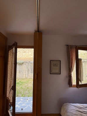 Ein Zimmer mit einem Bett, einem mit Vorhängen versehenen Fenster, einem Bild in einem Rahmen an der Wand und einer Steinsäule außen.