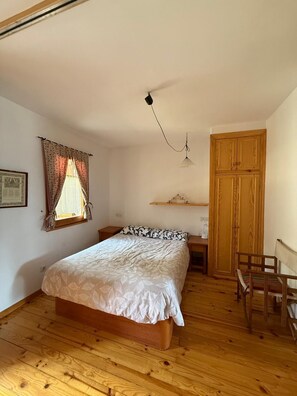 Une chambre avec un plancher en bois, un lit recouvert d’une courtepointe blanche, un bureau avec une chaise, une armoire en bois et une fenêtre avec des rideaux.