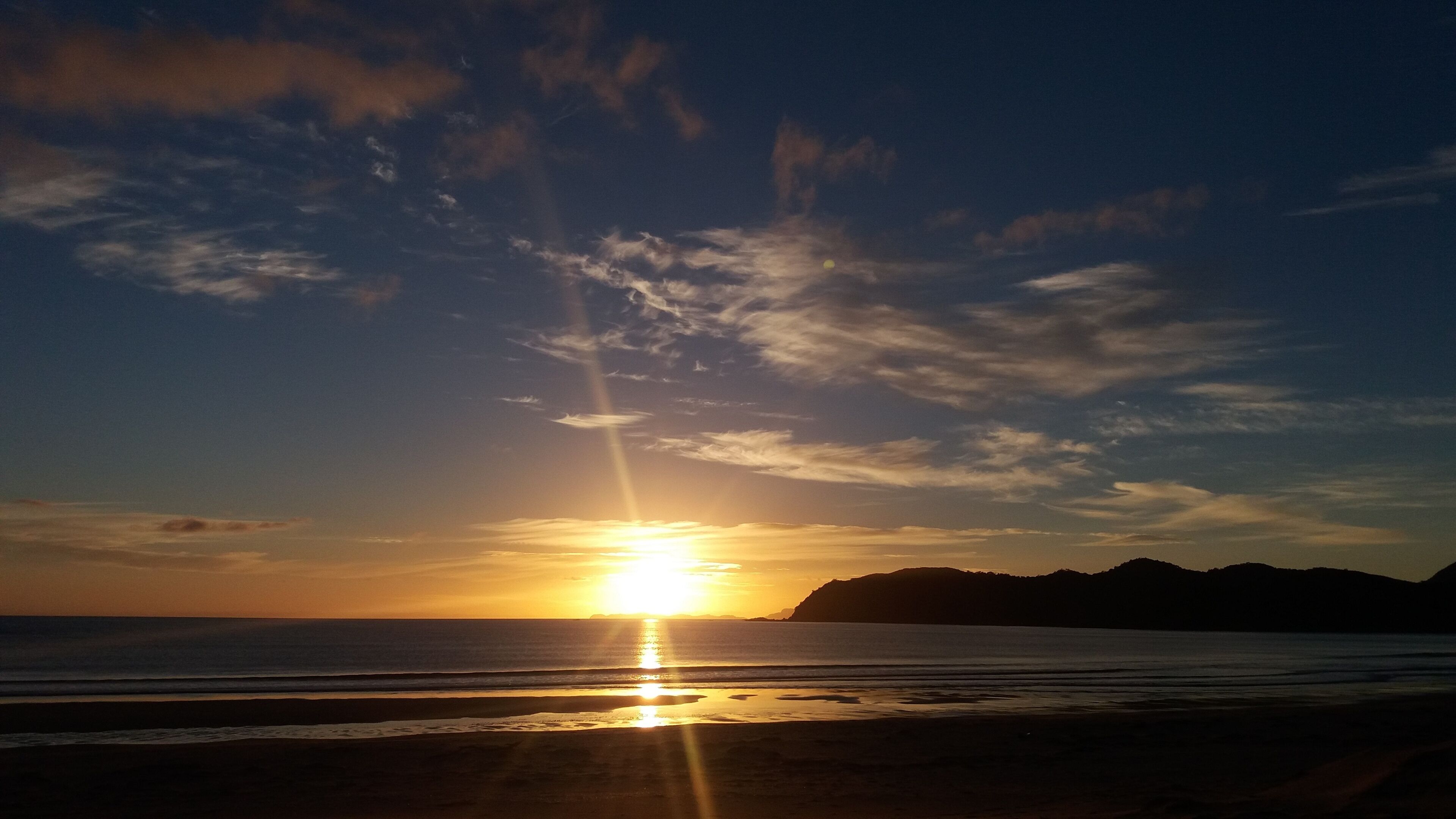 Un atardecer sobre una playa, con el sol bajo en el horizonte, proyectando un resplandor cálido sobre el agua y el cielo.