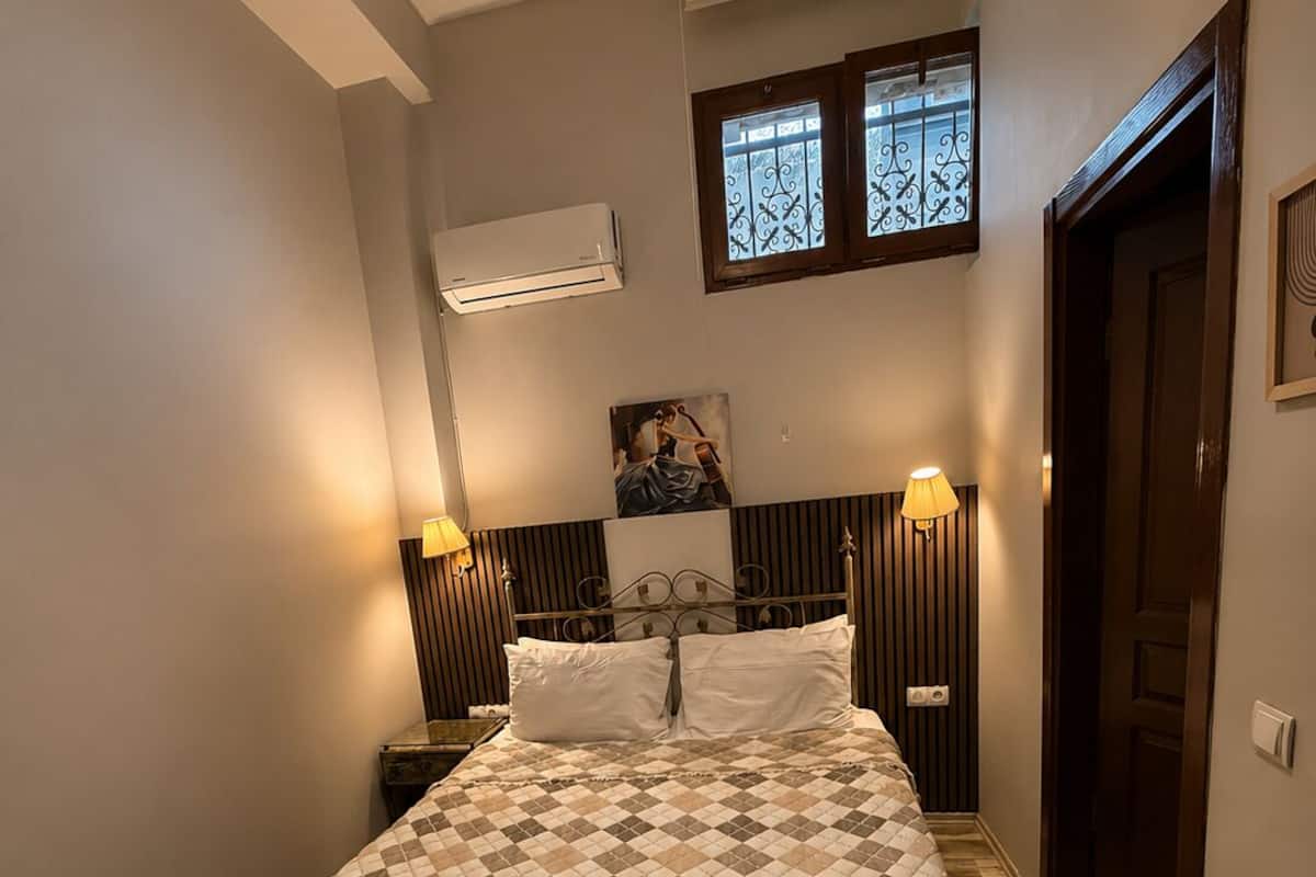 A bedroom with a bed, a chandelier, an air conditioning unit, a painting, and a door.