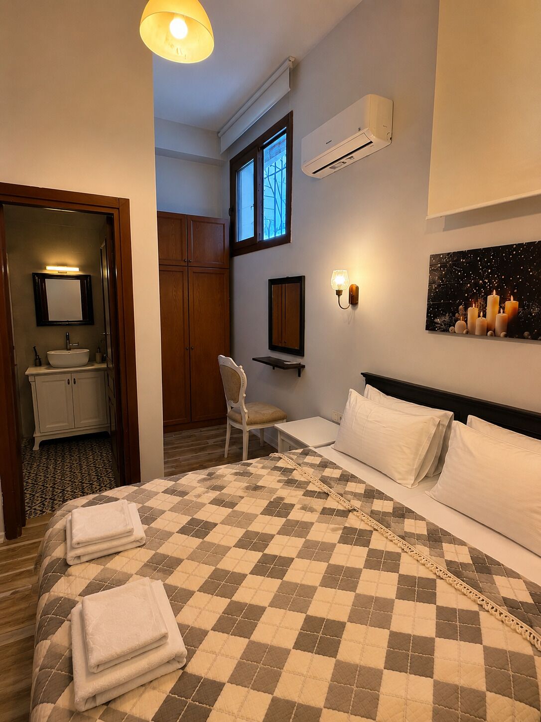A hotel room with a checkered bedspread, a bedside table, a chair, a window, an air conditioning unit, and a mirror.
