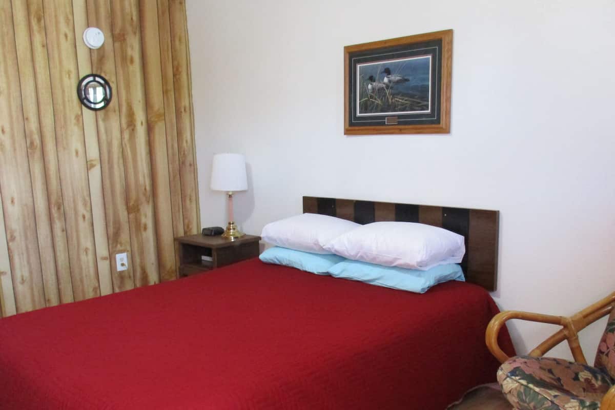 A room with a wooden panel wall, a bed with a red blanket, a nightstand with a lamp, and a framed picture on the wall.