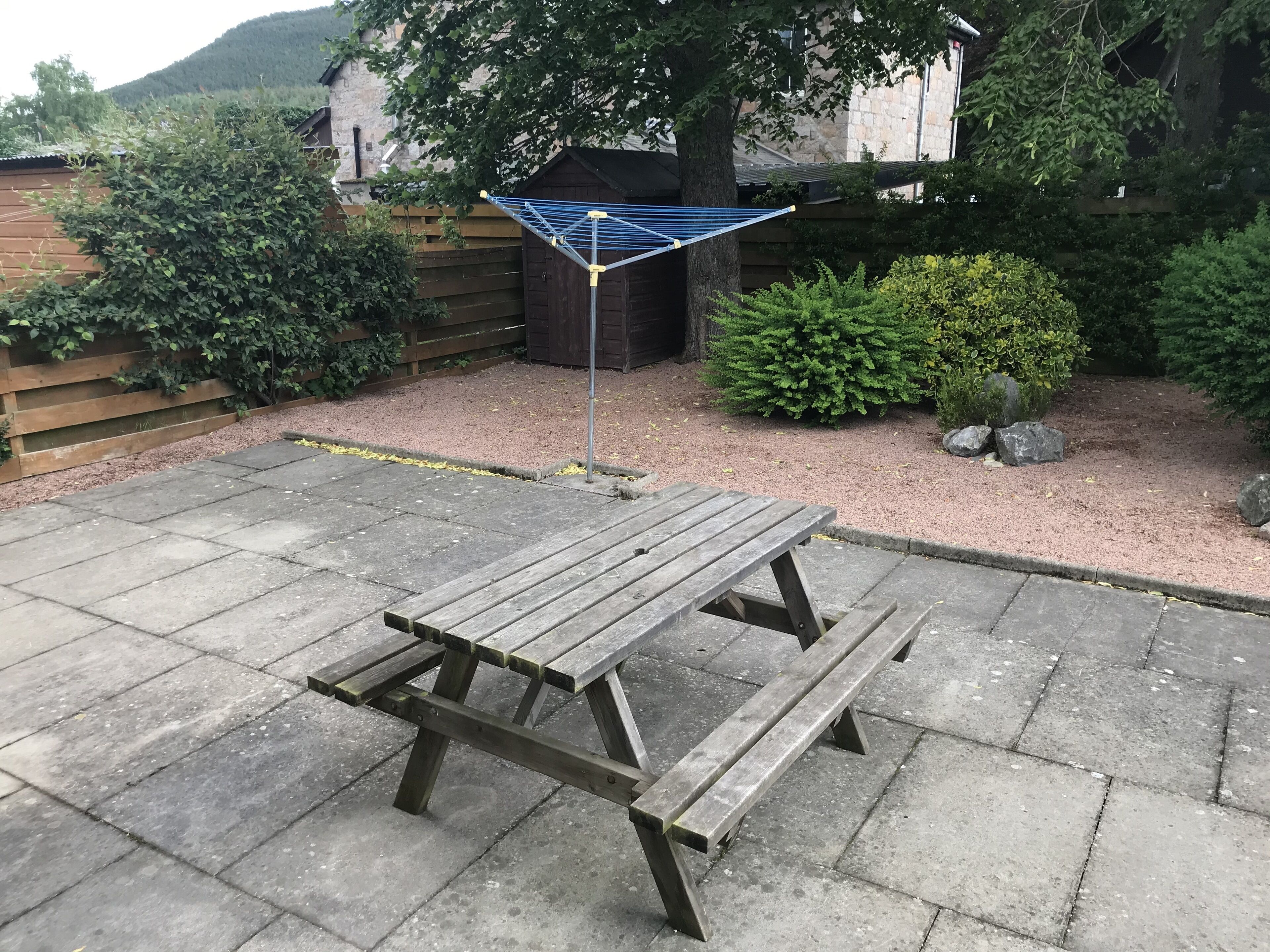 Una mesa de picnic de madera con estructura metálica, ubicada en un patio empedrado con una zona de grava y una valla de madera al fondo.
