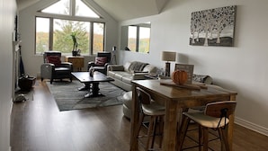 A spacious living room with a high ceiling, large windows, a wooden dining table, and a cozy seating area.