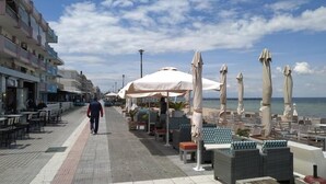 A seaside promenade with outdoor seating, a person walking, and a row of buildings along the water. - Alkyon Perea Apartments (Thermaikos)