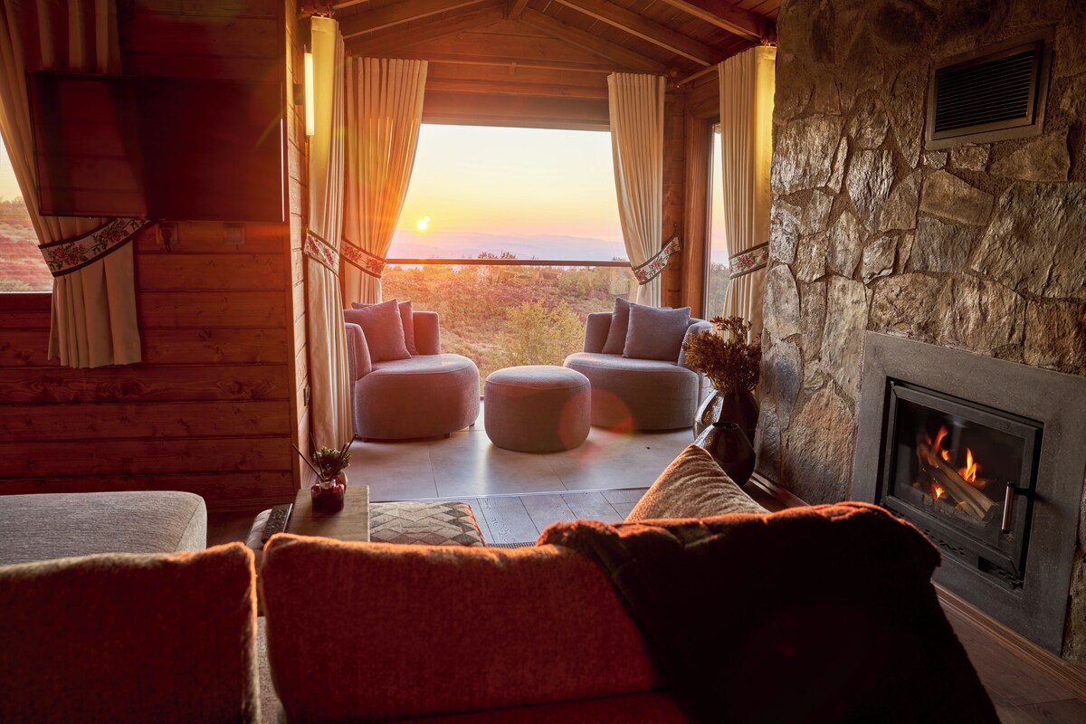 A cozy log cabin interior with a stone fireplace, plush seating, and a scenic view.