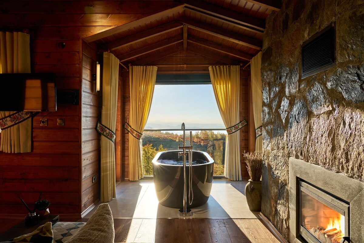 A wooden cabin interior with a stone fireplace, a large bathtub with a showerhead, and a view of the outdoors.