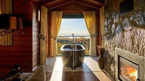 A wooden cabin interior with a stone fireplace, a large bathtub with a showerhead, and a view of the outdoors.