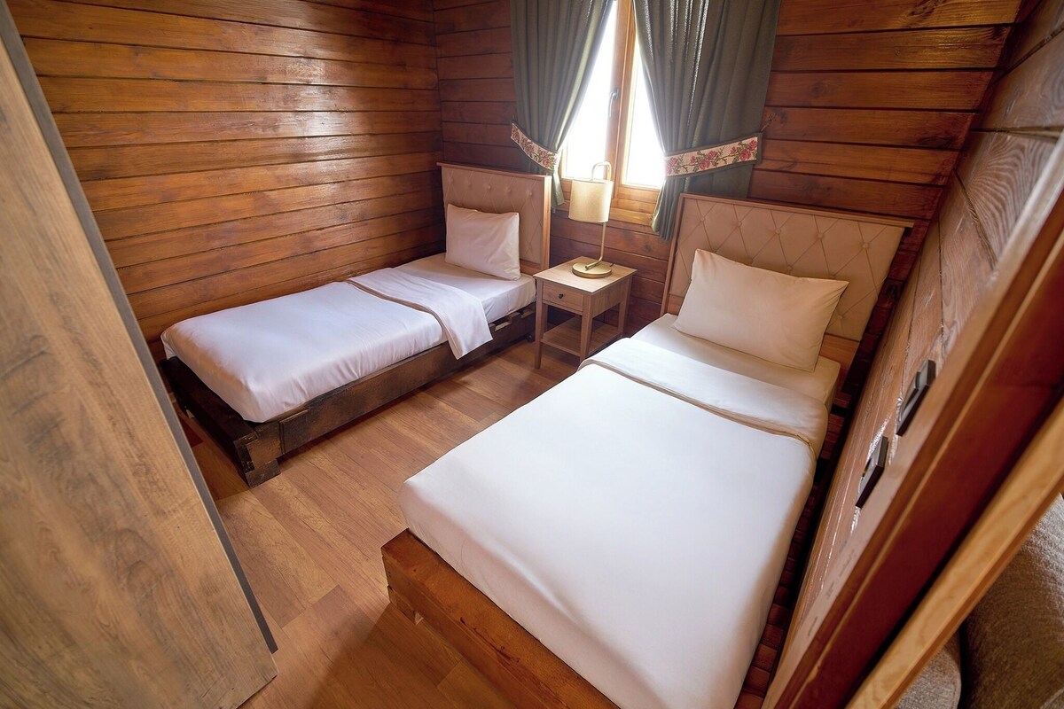 A wooden cabin room with two beds, a small table, and a window with curtains.