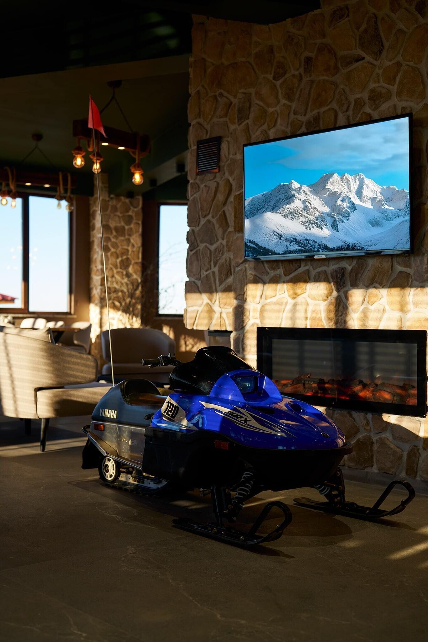 Une pièce avec un mur en pierre, un téléviseur fixé au mur qui diffuse une image de montagnes enneigées, et un motoneige Yamaha en premier plan.