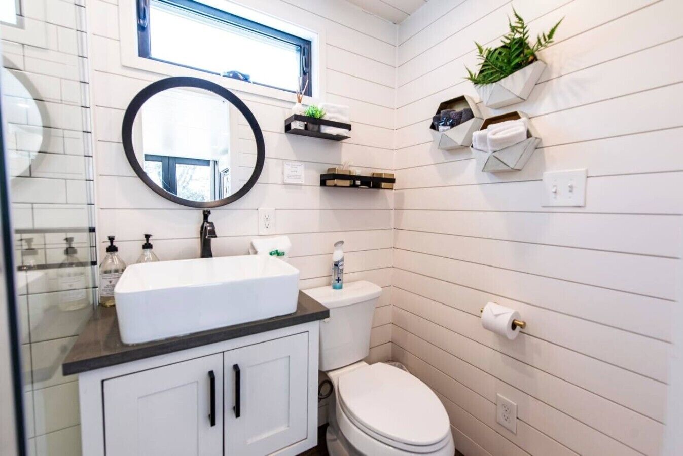 Une salle de bain avec un lavabo blanc, un plan de toilette noir, des toilettes blanches et un miroir. On y trouve des étagères avec des serviettes et une plante en pot.