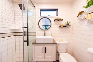 Un baño con cabina de ducha de vidrio, un lavabo blanco, un inodoro y una pequeña ventana sobre el lavabo.