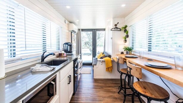A modern kitchen with a central island, a dining area with bar stools, and large windows with blinds. - Honeycomb Container Home | "Pickleball Court!" (Waco)