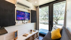 A modern living room with a flat-screen TV, a sofa with a yellow pillow, a small table with a chalkboard sign, and a view of the outdoors through a glass door. - Honeycomb Container Home | "Pickleball Court!" (Waco)