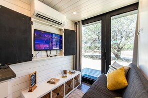 Una sala de estar moderna con un televisor de pantalla plana, un sofá con un cojín amarillo, una mesita con un cartel de pizarra y vistas al exterior a través de una puerta de cristal.